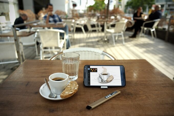 Tablet showing coffee drawing next to cup of coffee, biscuit, glass of water, and pen on café table