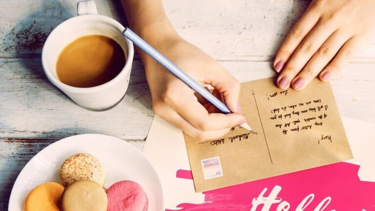 Hand writing a postcard with a blue pen next to coffee and colourful macarons on table