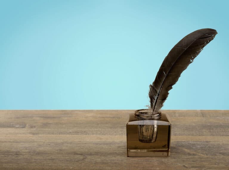 A feather quill pen resting in an inkwell on a wooden table, with a blue background