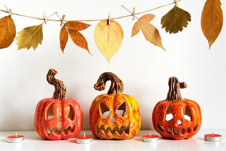 A row of three carved ceramic pumpkins with tealights, underneath a leaf garland — representing autumn and Halloween.