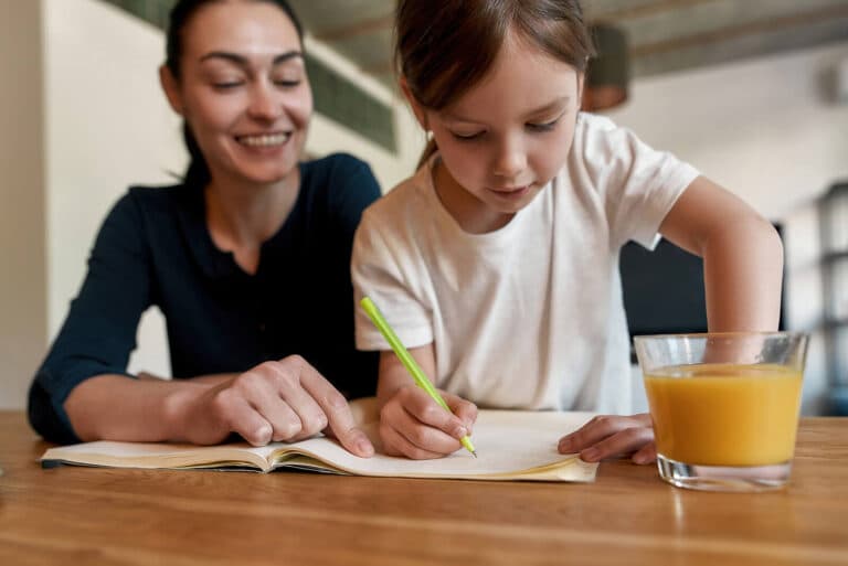Young girl writing in notebook with woman smiling beside her and glass of orange juice nearby