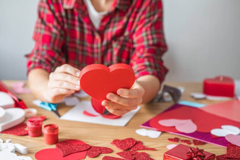 Person crafting red heart-shaped holiday card with paper and glue on wooden table