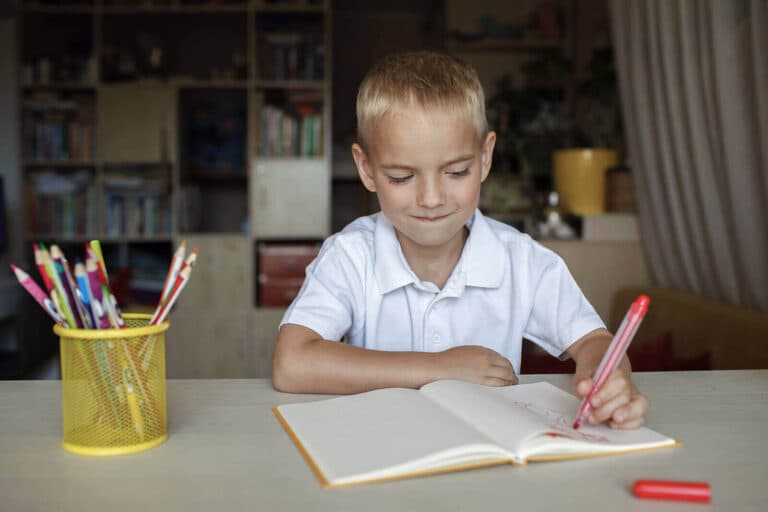 Young left-handed boy writing in notebook with red pen at table with pencils