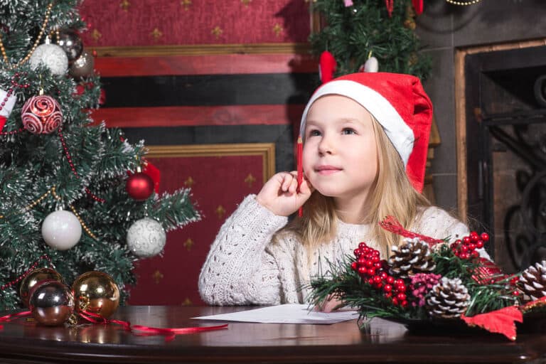A little girl writing a letter to Santa, wearing a Santa hat and sat next to a Christmas tree.