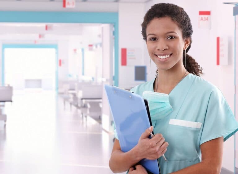 A young, smiling Black nurse wearing blue and stood in a hospital corridor, holding a clipboard and pen.