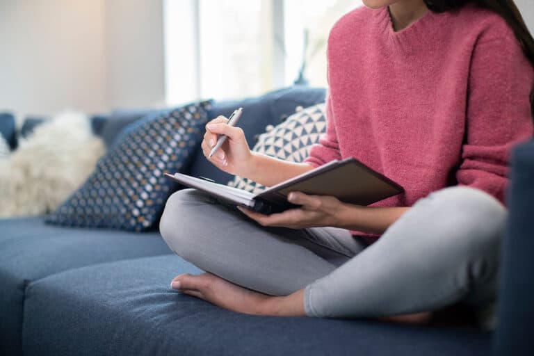 A close-up of a woman writing in a journal while sat on the sofa.