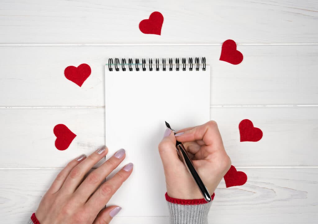 A Valentine's Day-themed image featuring a notepad, a hand holding a pen, and several red paper hearts.