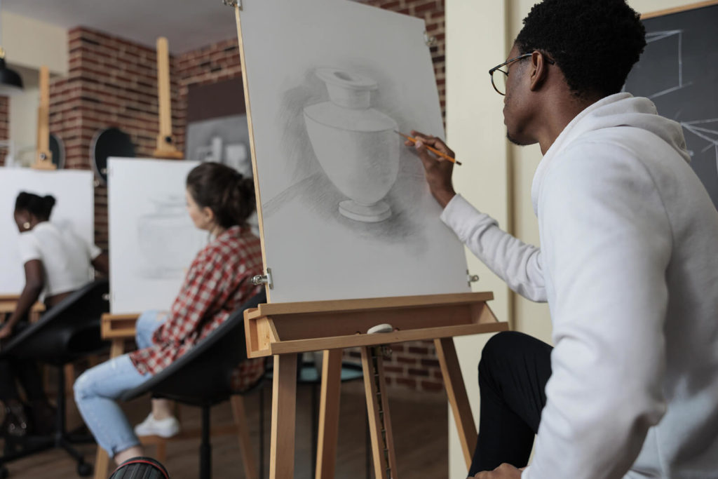 A Black art student wearing a white shirt, drawing a vase in pencil on an easel in an art class