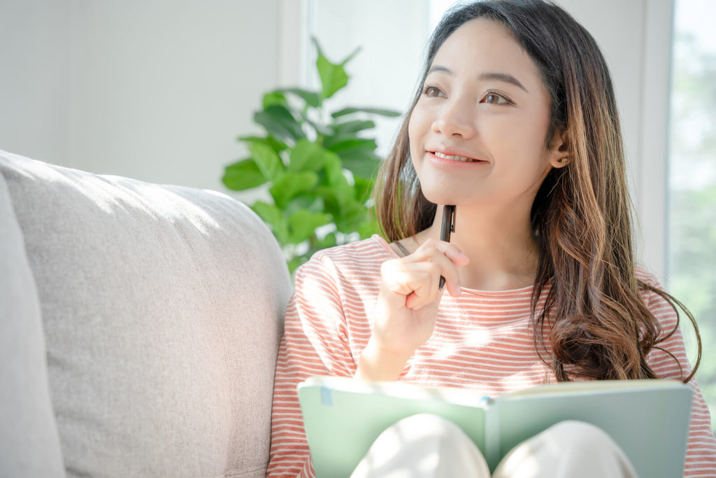 An Asian woman sat on a sofa, thinking about what to write or create in her art journal.