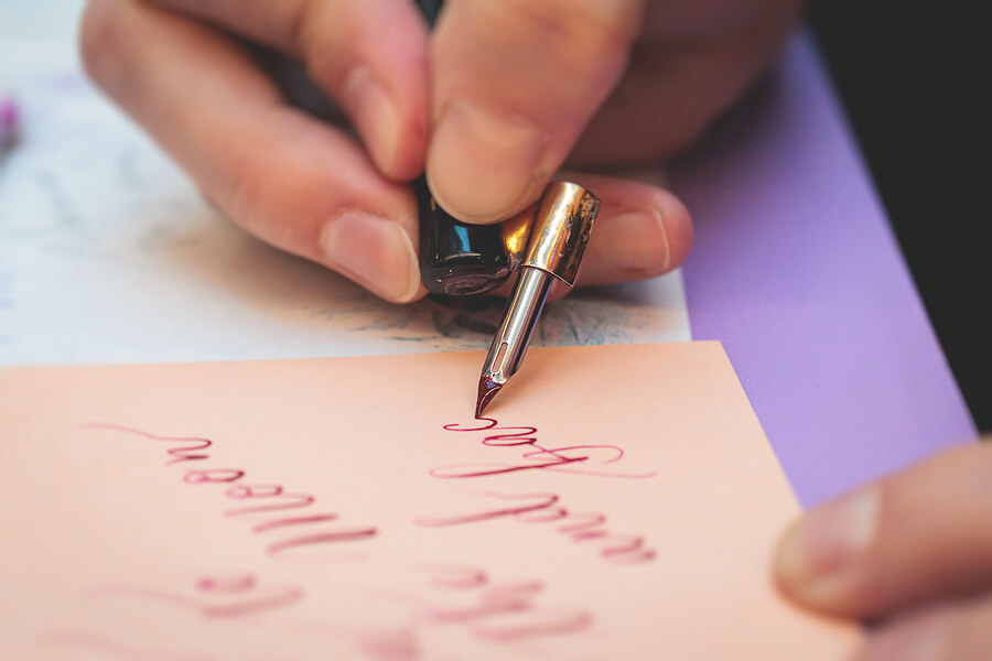 Process of calligraphy handwriting with an ink fountain pen feather, calligrapher practicing writing on postcard paper using pen brush and sign pen with inkwell on master-class lesson, hands view