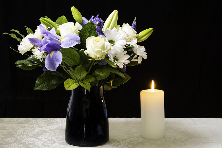 A black vase full of purple and cream blooms on a table next to a lit candle, representing a solemn occassion such as a funeral.