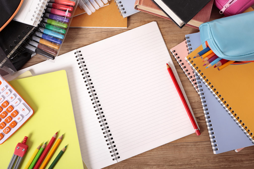 Busy student's desk with open notebook school bag text books and various pencils and crayons.