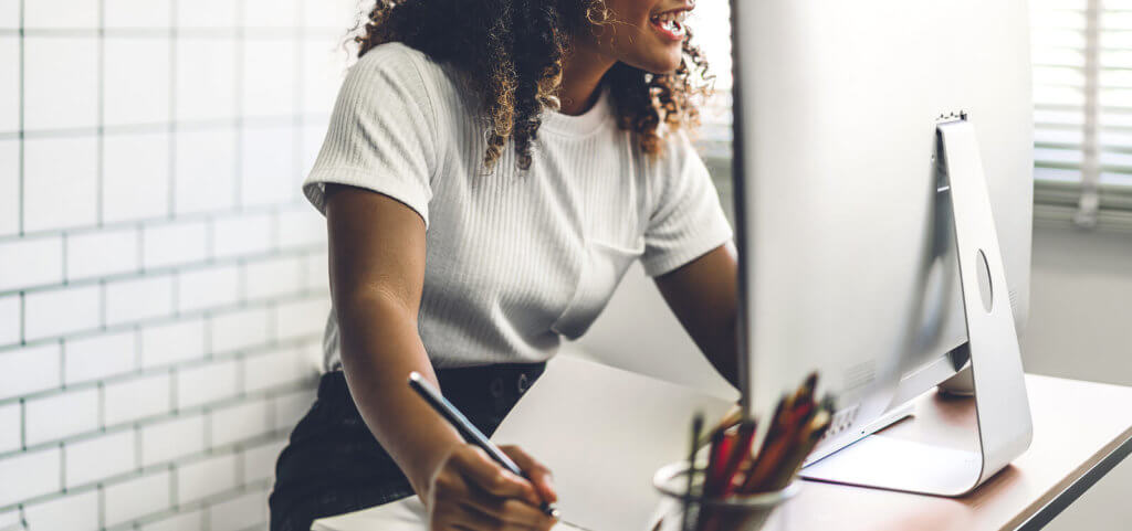 A woman writing on a notepad while laughing at a laptop screen.