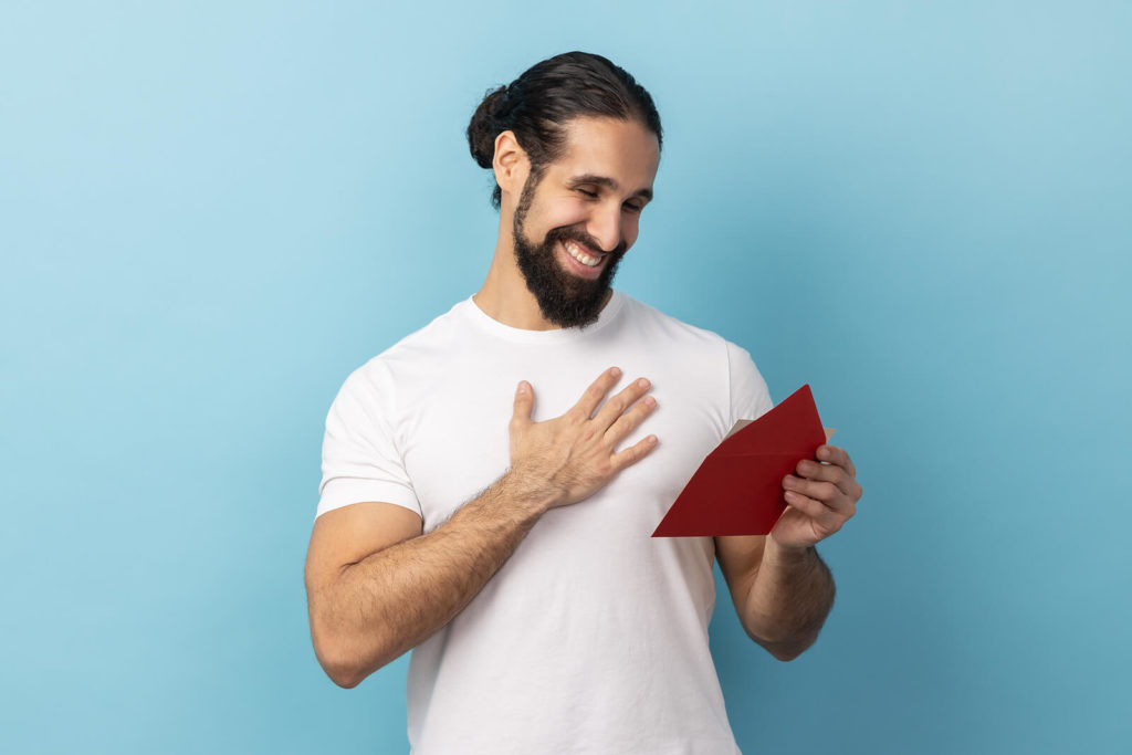 A man in a white t-shirt reading a love letter, clutching his chest and smiling.