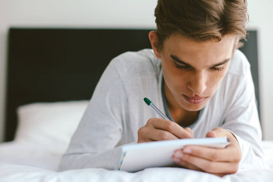 teenage boy writing story on bed with pen and paper