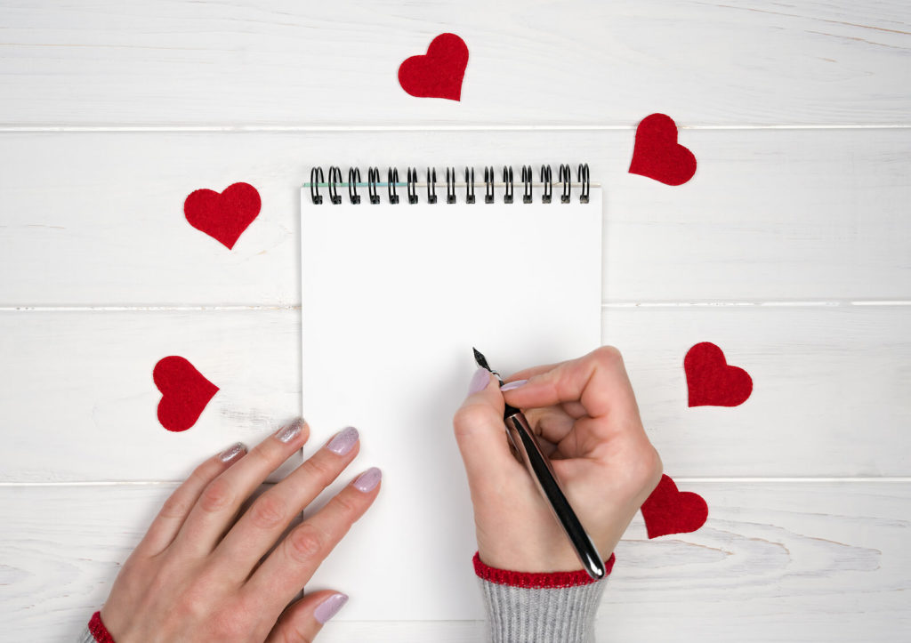 A Valentine's Day-themed image featuring a notepad, a hand holding a pen, and several red paper hearts.