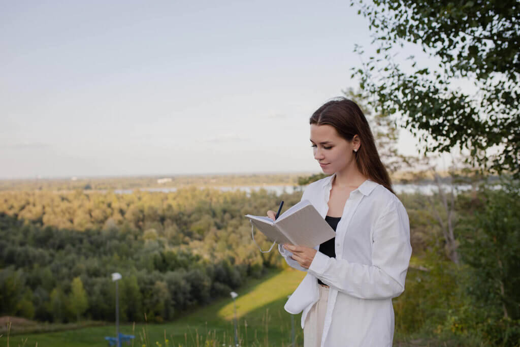 A woman stood in the countryside wearing a white shirt and writing in a book