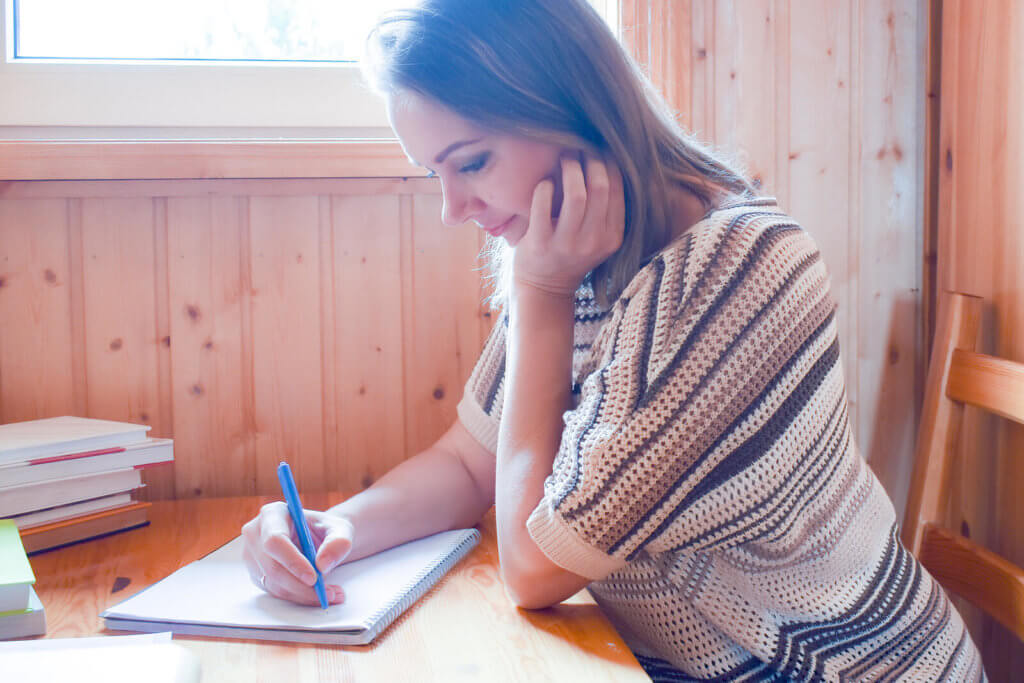 woman writing at desk in a notepad, sat next to a window