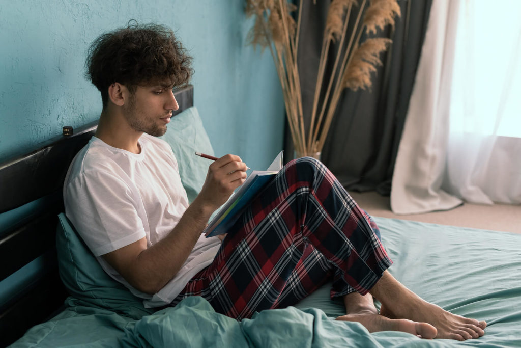 A young man writing a diary while sat on his bed wearing pyjamas.