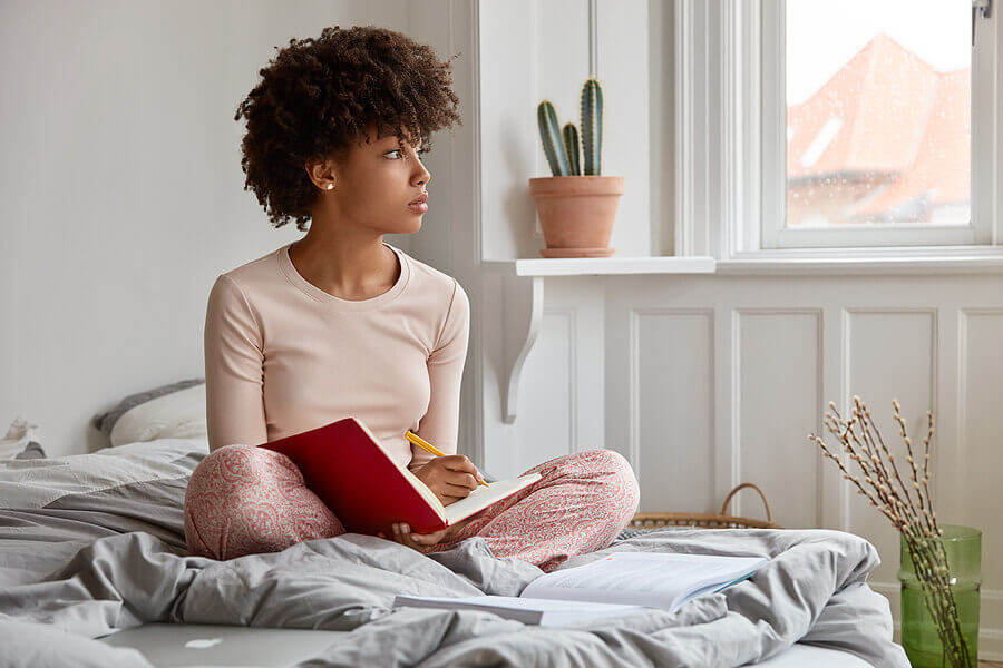 young woman sat on bed, writing in notepad and looking thoughtful